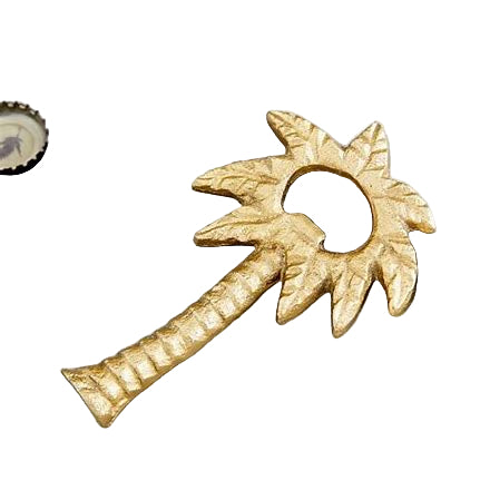 A brass bottle opener shaped like a palm tree, with gold finish, placed on a grey surface next to a glass bottle and a bottle cap.